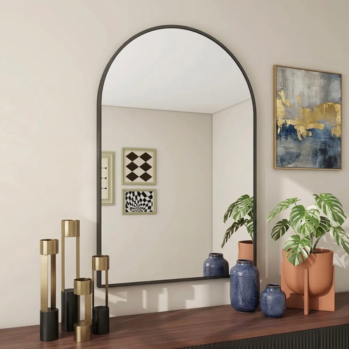 Large arch-shaped mirror on a wooden surface with decorative vases and plants in a room.