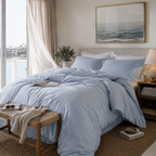 Neatly made bed with light blue cotton bedding in a bedroom with large windows and a bench.