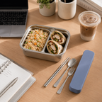 Lunchbox with food, utensils, and a drink on a wooden table.