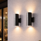 Two outdoor wall lights on a concrete wall with plants and a window in the background.